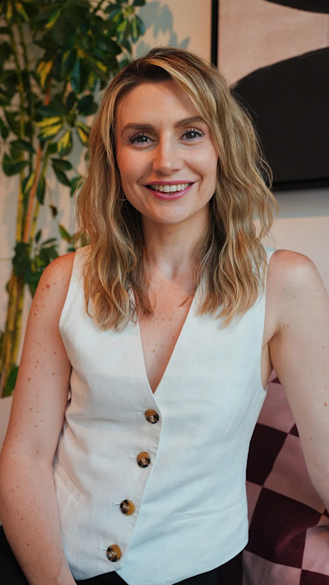 Smiling woman with shoulder-length blonde wavy hair wearing a white vest, seated indoors at Palma Hair Concept salon.